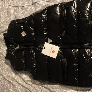 Säljer en snygg svart dunväst från Moncler i storlek M. Västen har en glansig finish och är perfekt för kyliga dagar. Den har dragkedja framtill och två praktiska fickor med dragkedjor. Moncler-loggan sitter på bröstet. Perfekt för både vardag och fest!