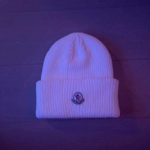 Vit ribbad mössa från Moncler - Säljer en snygg vit ribbad mössa från Moncler. Den är i nytt skick och har en broderad logga framtill. Perfekt för vintern och håller dig varm och stilren. Passar de flesta tack vare one size-storleken.