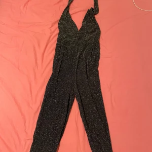 Svart glittrig jumpsuit - Snygg svart glittrig jumpsuit perfekt för festliga tillfällen. Den har en djup v-ringning och halterneck-design som ger en elegant look. Materialet är stretchigt och bekvämt, vilket gör den lätt att bära hela kvällen. Perfekt för nyår eller andra speciella tillfällen! Priset kan alltid diskuteras! XS/S och har endast använts 1-2 gånger!