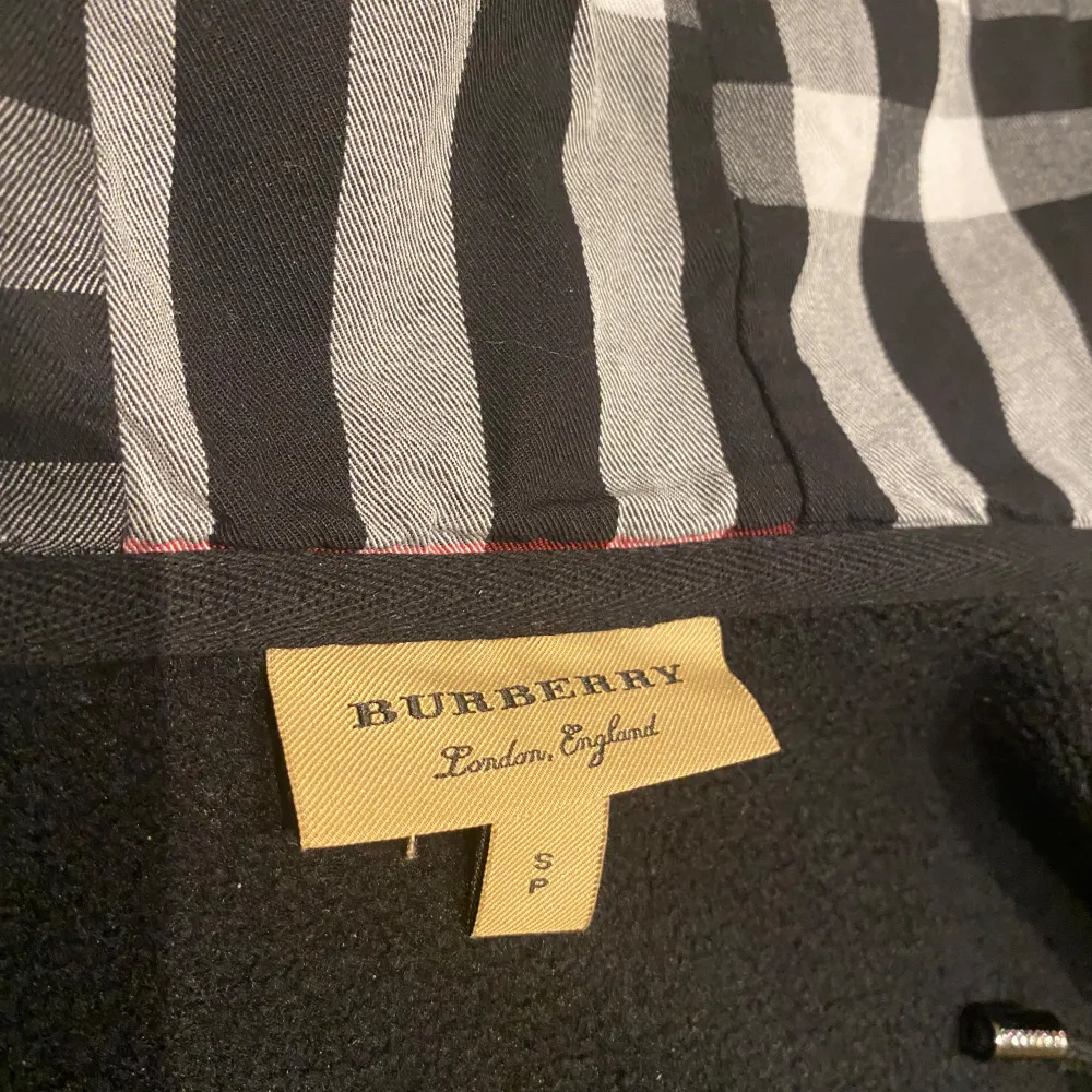 Säljer en snygg svart hoodie från Burberry i storlek S. Den har ett klassiskt rutigt foder i huvan med grå, vita och röda detaljer. Dragkedja framtill och broderad logga på bröstet. Perfekt för höst och vinter!. Neuletakit & Villapaidat.