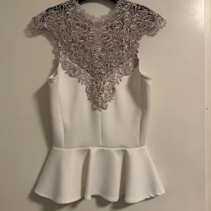Vit topp med spetsdetaljer - Säljer en elegant vit topp med vacker spetsdetalj i halsen. Toppen är ärmlös och har en volangkant nedtill som ger en fin siluett. Perfekt för festliga tillfällen eller en utekväll. Den är i mycket bra skick och redo att bäras av någon som vill sticka ut.