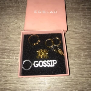 Smyckeset från Edblad  - Säljer ett coolt smyckeset från Edblad. Setet innehåller flera ringar i guld och silver, inklusive en med texten 'GOSSIP'. Perfekt för att ge din outfit en edgy touch. Kommer i en snygg rosa ask.