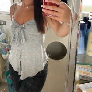 Grå ärmlös topp med knyt - Säljer en superfin grå ärmlös topp med knyt framtill. Perfekt för sommardagar eller en avslappnad fest. Den är i mjukt material och har en lös passform som gör den bekväm att bära hela dagen.