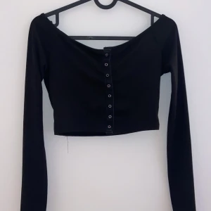 Svart ribbad offshoulder topp - Snygg svart ribbad offshoulder topp med knappar framtill. Perfekt för en kväll ute eller en avslappnad dag. Den är croppad och har långa ärmar, vilket gör den både trendig och bekväm. Passar bra till både jeans och kjol!