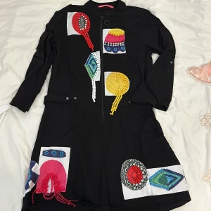 Svart klänning med färgglada detaljer - Säljer en unik svart klänning från Desigual med coola, färgglada applikationer i rött, gult och blått. Klänningen har en dragkedja framtill och långa ärmar. Perfekt för dig som vill sticka ut på festen!