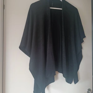 Svart glittrig kavaj från Å - Säljer en elegant svart poncho med glittriga detaljer från märket Å frpn Åhlens. Perfekt för festliga tillfällen med sin stilrena design och glittriga yta. Den är både bekväm och chic.