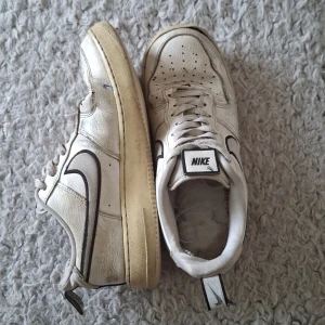 Vita Nike Air Force sneakers - Säljer ett par vita Nike Air Force sneakers i storlek 42.5. Skorna är välanvända med synliga tecken på slitage, särskilt på sulorna som har gulnat. De har en klassisk design med svart Nike-logga på sidan och snörning framtill. Perfekta för någon som gillar en vintage look eller vill ha ett par bekväma vardagsskor.