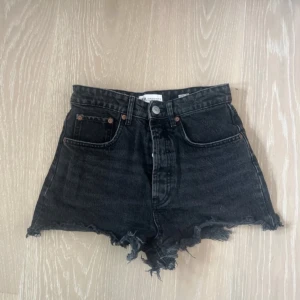Svarta jeansshorts från Zara - Säljer ett par svarta jeansshorts från Zara i storlek 36. De är högmidjade och har en snygg, sliten look med fransiga kanter. Perfekta för sommaren eller en avslappnad dag ute. De har knappar framtill.