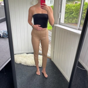 Beige skinnbyxor från Zara - Beige ”skinnbyxor” från Zara. Högmidjade med dragkedja i sidan. Slits med dragkedja vid benslut. Byxorna är i syntetmaterial. Defekt i midjan (syns ej med plagget på, se bild).