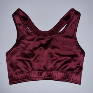 Vinröd sporttopp från Newbody - Säljer en snygg och bekväm vinröd sporttopp från Newbody. Toppen är tillverkad i ett glansigt material som känns mjukt mot huden. Perfekt för träning eller andra fysiska aktiviteter. Den har en tight passform och breda axelband för extra stöd.