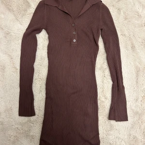Brun ribbad långärmad klänning  - En brun ribbad långärmad klänning med slits vid ärmarna. Den har en snygg krage och knappar vid brösten