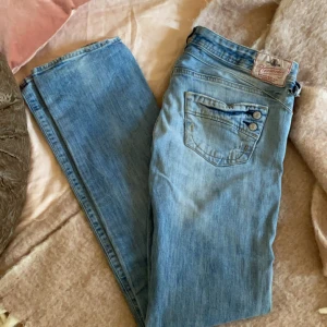 Snygga raka jeans med låg midja i ljusblå denim -  Dessa trendiga jeans i jusblå denim med låg midja har en tidlös look. Jeansen har en stilren design med tre knappar på den bakre fickan för en unik touch. Perfekt för vardags och festtillfällen.   Storlek: 29/34 100% bomull Mycket bra skick!  