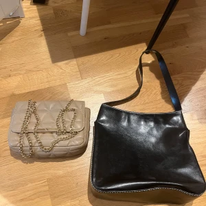 Beige quiltad handväska - Säljer en superfin beige handväska med quiltat mönster. Den har en elegant kedja som axelrem och en praktisk knappstängning. Perfekt för både vardag och fest! Säljer även en svart väska med vita sömmar.