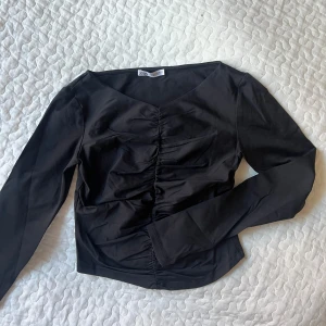 Svart långärmad topp från Zara - Snygg svart långärmad topp från Zara med rynkdetaljer framtill. Perfekt för en stilren look. Toppen har en elegant v-ringning och är tillverkad i ett mjukt material.
