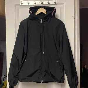 Svart vindjacka från Moncler - Säljer en stilren svart vindjacka från Moncler med dragkedja och hög krage. Jackan har långa ärmar och Moncler-logga på armen. Perfekt för blåsiga dagar.