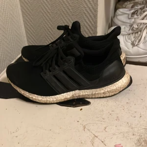 Svarta Adidas Ultraboost - Säljer ett par svarta Adidas Ultraboost med snörning och en bekväm sula. Skorna har en stilren design med de klassiska tre ränderna på sidan. Perfekta för både träning och vardag.