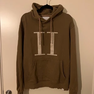 Brun hoodie från Les Deux - Säljer en snygg brun hoodie från Les Deux med en stor logotyp framtill. Hoodien har en klassisk känguruficka och justerbar huva med dragsko. Perfekt för en avslappnad stil.