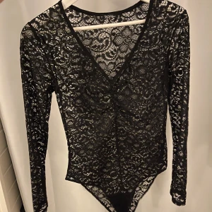 Svart spetsbody med v-ringning - Snygg svart spetsbody med v-ringning och långa ärmar. Perfekt för en kväll ute eller en speciell tillställning. Den genomskinliga spetsen ger en elegant och stilren look.