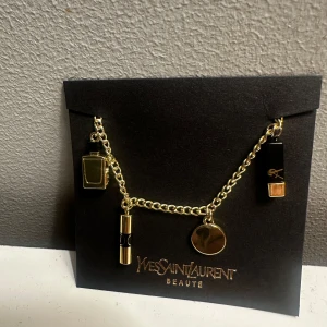 Guldarmband med berlocker från Yves Saint Laurent - Elegant guldarmband från Yves Saint Laurent med flera berlocker, inklusive en parfymflaska, ett läppstift och en rund medaljong. Perfekt för att lägga till en lyxig touch till din stil.