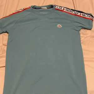 Blå t-shirt från Moncler - Snygg blå t-shirt från Moncler med korta ärmar och logotypdetaljer på axlarna. T-shirten har en rund halsringning och är tillverkad i ett mjukt material för extra komfort. Qr kod funkar. Du vet varför den är billig så fråga inte