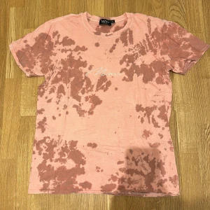 Rosa t-shirt från MAN - Säljer en rosa t-shirt från MAN med ett unikt batikmönster. T-shirten har korta ärmar och ett tryck med texten 'Man' på bröstet. Perfekt för en avslappnad stil.