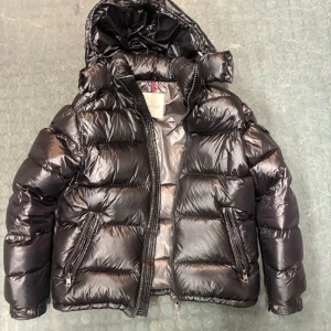 Moncler maya  - Säljer en snygg svart pufferjacka från Moncler. Jackan har en glansig finish och är utrustad med en huva och dragkedja framtill. Den har också praktiska fickor med dragkedjor och en ikonisk Moncler-logga på ärmen. Perfekt för kalla dagar! Köpt för 4500kr dustbag medföljer och även tags👍🏼