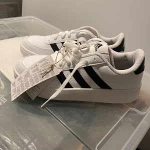 Vita sneakers från Adidas - Snygga vita sneakers från Adidas med klassiska svarta ränder. Skorna har en stilren design med snörning och är perfekta för en sportig look. De är märkta med Adidas-loggan på plösen. Aldrig använda och lappen sitter kvar.