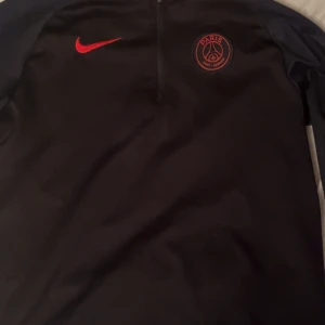 Svart långärmad tröja från Nike - Säljer en svart långärmad tröja från Nike med Paris Saint-Germain-logga på bröstet. Tröjan har en dragkedja vid halsen och är perfekt för fotbollsträning eller vardagsbruk. Den har en stilren design med röda detaljer.