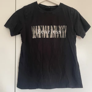 Svart t-shirt med tryck - Snygg svart t-shirt med texten 'Fearless' i ett zebramönstrat tryck på bröstet. Perfekt för en cool och avslappnad stil. Kortärmad och i mjukt material.