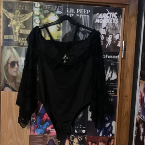 Svart offshoulder topp med spetsdetaljer - Snygg svart offshoulder topp med spetsdetaljer och en dekorativ korsdetalj framtill. Toppen har långa spetsärmar som ger en elegant look. Perfekt för en unik stil.