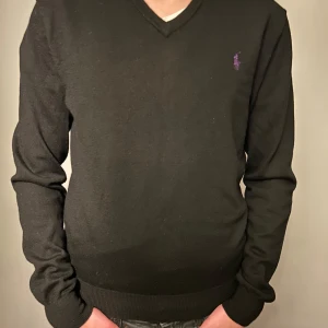 Svart tröja med lila logga - Snygg svart tröja från Ralph Lauren med en lila broderad logga på bröstet. Tröjan har en v-ringad design och långa ärmar, perfekt för en stilren look. Passar bra till både jeans och chinos.