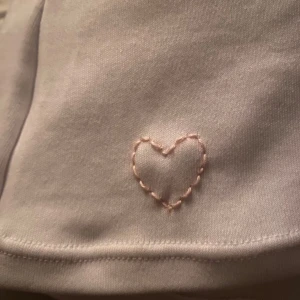 Vit kortärmad t-shirt med hjärtdetalj - Säljer en söt vit kortärmad t-shirt med ett litet broderat hjärta i ljusrosa. Toppen är i ett mjukt material och har en ribbad kant vid halsen.💕