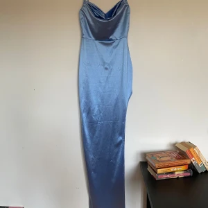 Ljusblå långklänning från Windsor - Elegant ljusblå långklänning från Windsor i satin. Klänningen har en vacker draperad halsringning och en figurnära passform med en slit på ena benet som ger en stilren siluett.  Perfekt för speciella tillfällen där du vill känna dig extra glamorös.