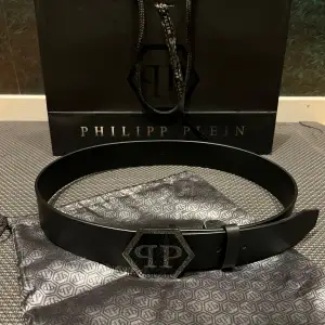 Snyggt svart läder bälte(100% kalvskinn) från Philipp Plein med en unik hexagonformad spänne i silver. Tillverkat i Italien,  bältet är perfekt för att ge en stilren touch till din outfit och ger en känsla av lyx. Bältet har en elegant design med märkets logotyp ingraverad på spännet som blänker. Köpt på ”Plaza Major” i Spanien i Phillip Pleins butik( ordinarie pris 5500kr) Säljer då jag tyvärr växt ur den. Pris kan diskuteras lite vid snabb affär