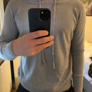Grå hoodie med dragsko - Säljer en stilren grå hoodie med dragsko i huvan. Perfekt för en avslappnad look. Tröjan har långa ärmar och en mjuk känsla.