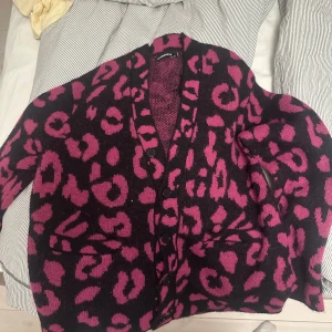 Svart och rosa leopardmönstrad kofta från Lindeberg - Säljer en svart och rosa leopardmönstrad kofta med knappar framtill. Koftan har en mysig och varm känsla, perfekt för kyligare dagar. Den har långa ärmar och en avslappnad passform. Storlek L funkar som M