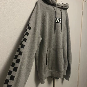 Grå hoodie från Vans - Säljer en snygg grå hoodie från Vans i storlek small. Den har en klassisk logga på bröstet och fina rutiga detaljer på ärmen. Väldigt skön och behagligt hoodie som tyvärr inte kommer till användning. Superbekväm och i bra skick!