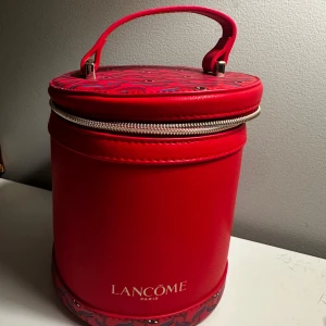 Röd rund väska från Lancôme - Snygg och unik rund väska från Lancôme i en livfull röd färg. Väskan har ett praktiskt handtag och en dragkedja som går runt hela toppen. Insidan är mönstrad med ett coolt designmönster. Perfekt för att förvara dina skönhetsprodukter eller som en statement-accessoar.