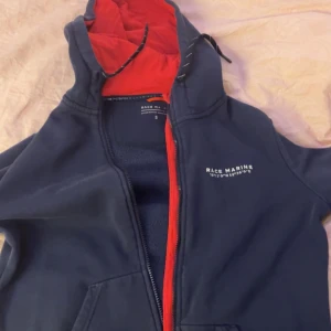 Mörkblå hoodie från Race Marine - Säljer en mörkblå hoodie från Race Marine med röd insida i huvan och dragkedja. Den har en sportig look med tryck på bröstet och är perfekt för kyliga dagar. Huvan har justerbara snören för extra komfort.