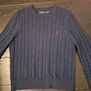Mörkblå stickad tröja från Polo Ralph Lauren - Säljer en snygg mörkblå stickad tröja från Polo Ralph Lauren med kabelstickat mönster och röd broderad logga på bröstet. Tröjan har långa ärmar och ribbade kanter. Perfekt för kyligare dagar! Modellen är i M men sitter som en S eftersom den krympt i tveten. Super bra kvalitet och har inga skador! Använd bara några gånger. Ny pris ca 2000kr!