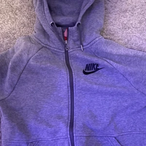 Grå hoodie från Nike - Säljer en grå hoodie från Nike i bra skick. Den har en dragkedja framtill och en klassisk Nike-logga på bröstet. Perfekt för både träning och vardag. Den är mjuk och bekväm med en justerbar huva.