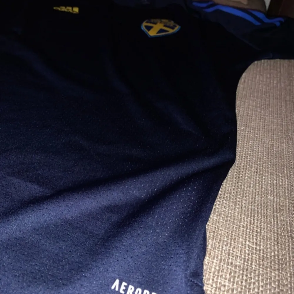 Säljer en snygg svensk landslagströja från Adidas i mörkblått med gula och blå detaljer. Tröjan har korta ärmar och är i AEROREADY-material för bra andningsförmåga. Perfekt för fotbollsfans som vill visa sitt stöd för Sverige!. T-shirts.