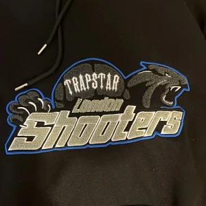 Svart hoodie från Trapstar - Säljer en svart hoodie från Trapstar med ett coolt 'London Shooters'-tryck på framsidan. Den har en stor ficka fram och justerbar huva med snören. Perfekt för en avslappnad stil eller streetwear-look.