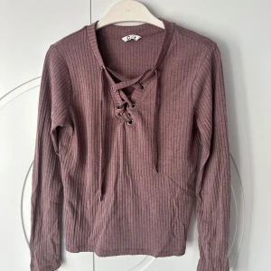 Lila ribbad tröja med snörning - Säljer en superfin lila ribbad tröja i storlek M. Den har långa ärmar och en snygg snörning framtill som ger en extra detalj. Perfekt för höst och vår när man vill vara både bekväm och stilren. Passar till både jeans och kjol!