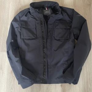 Svart jacka från Dressmann - stilren fieldjacker i marinblå färg från Dressmann. Den har långa ärmar och praktiska fickor framtill. Jackan är perfekt för höst och vår, med en dragkedja och knappar för extra säkerhet. har luva i kragen, sitter bra på dig 175-185cm, som ny!