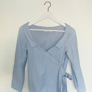 Omlottblus - En jättesnygg blus i ljusblå skjorttyg. Snygg knytdetalj och v-ringning. Sitter jättesnyggt på. 8/10 skick. 