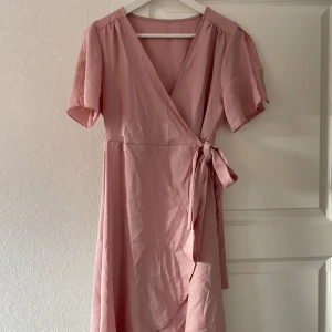 Rosa omlottklänning - Säljer en superfin rosa omlottklänning med korta ärmar. Den har en v-ringad design och knytning i midjan som ger en smickrande passform. Perfekt för sommarens alla fester eller en mysig vårdag. Klänningen är i ett mjukt och bekvämt material.