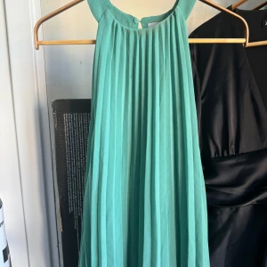 Grön klänning från H&M - Säljer en superfin grön klänning från H&M. Den har en luftig och plisserad design som ger en härlig rörelse. Perfekt för sommarens alla fester eller en dag på stan. Blusen är ärmlös och har en knappdetalj vid halsen.