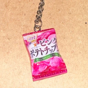 Nyckelring med rosa chipsförpackning - Ett supercoolt halsband som ser ut som en liten rosa chipsförpackning. Den har japansk text och en kedja i silverfärg. Perfekt för att ge din vardag lite extra stil!