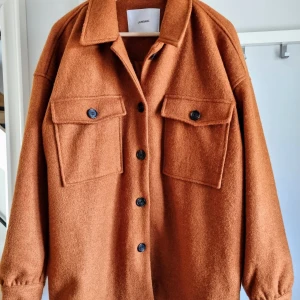 Brun overshirt från Junkyard - Säljer en snygg brun overshirt från Junkyard. Den har en cool och avslappnad stil med stora fickor framtill och knappar hela vägen. Perfekt för höst och vår när du vill ha något lätt men ändå varmt. Materialet känns mjukt och skönt, och den har långa ärmar med knappar vid ärmsluten.